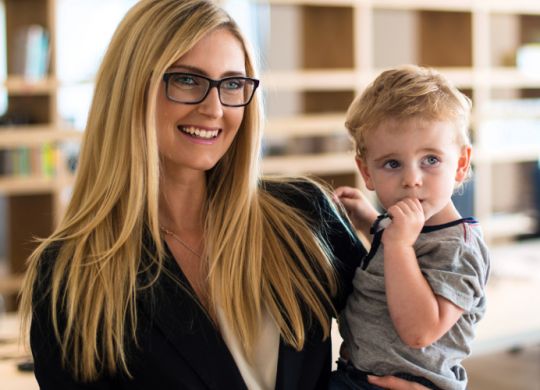 Reconciliation of family and work life: Attractive blond woman in business attire proudly carrying a small boy in her arm in office environment