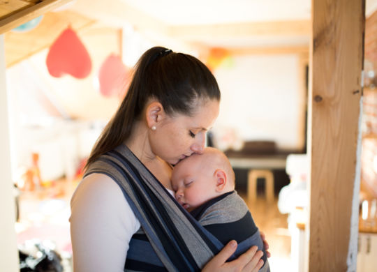 Beautiful young mother at home with her baby son sleeping in sling, kissing him on head