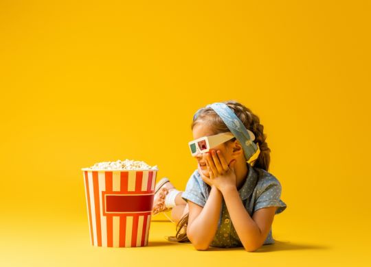happy kid in 3d glasses lying and looking at popcorn bucket on yellow
